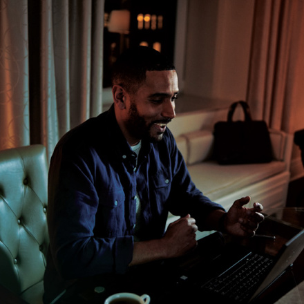 A man with a beard is sitting at a table in a dimly lit room, engaged in a video call on his laptop. A glass of water and a cup are on the table. The background shows a sofa, curtains, and a decorative wall.