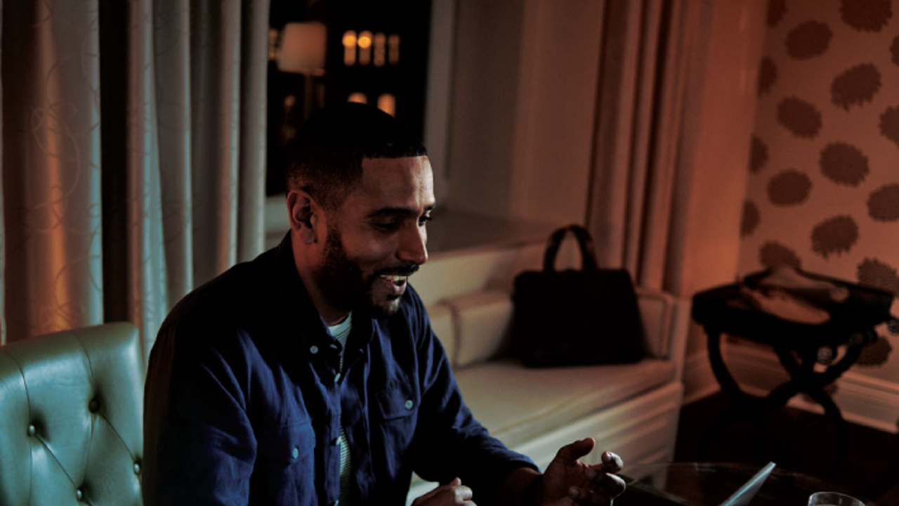 A man with a beard is sitting at a table in a dimly lit room, engaged in a video call on his laptop. A glass of water and a cup are on the table. The background shows a sofa, curtains, and a decorative wall.