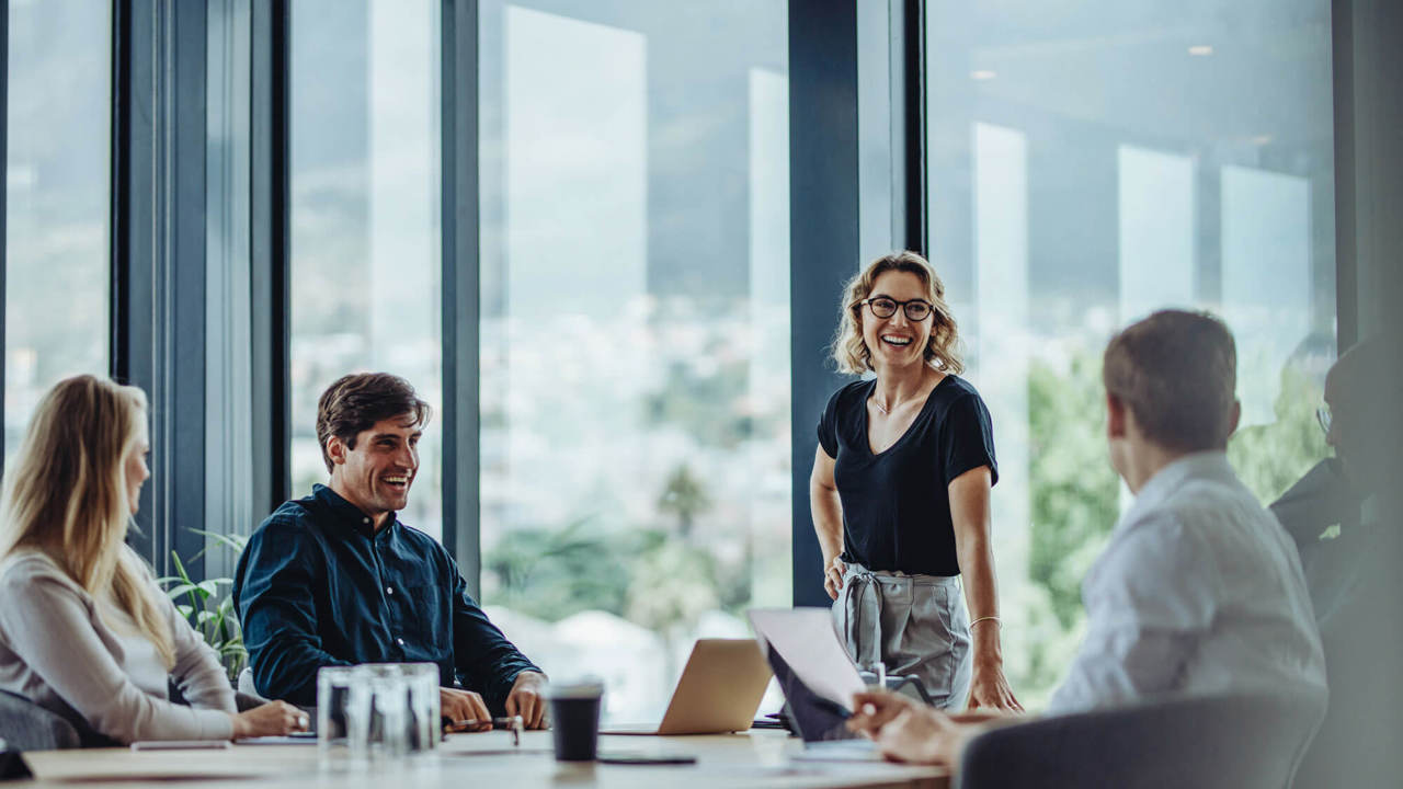 A group of people in a modern office setting, sitting and standing around a table. One person is standing and smiling, appearing to lead a discussion. Large windows provide a view of a cityscape outside.