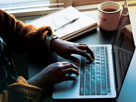 A person wearing a smartwatch types on a laptop. A notepad and a pen are nearby, and a coffee cup sits to the right. Sunlight filters through the blinds in the background, creating a cozy workspace atmosphere.