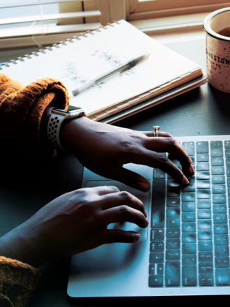 A person wearing a smartwatch types on a laptop. A notepad and a pen are nearby, and a coffee cup sits to the right. Sunlight filters through the blinds in the background, creating a cozy workspace atmosphere.