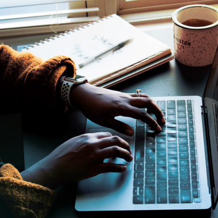 A person wearing a smartwatch types on a laptop. A notepad and a pen are nearby, and a coffee cup sits to the right. Sunlight filters through the blinds in the background, creating a cozy workspace atmosphere.