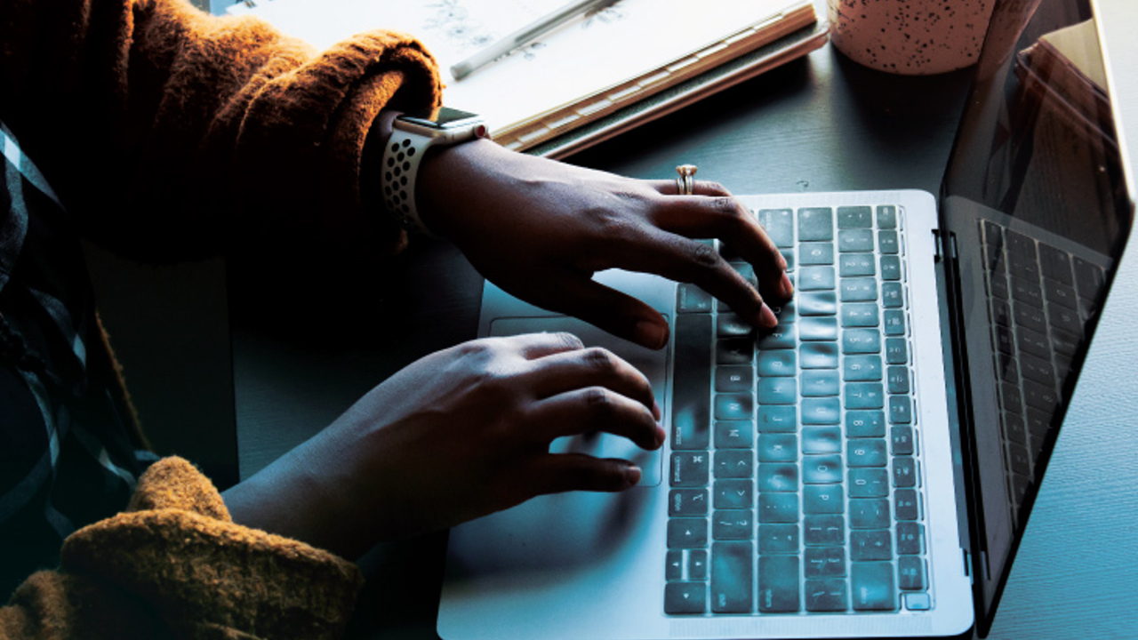 A person wearing a smartwatch types on a laptop. A notepad and a pen are nearby, and a coffee cup sits to the right. Sunlight filters through the blinds in the background, creating a cozy workspace atmosphere.