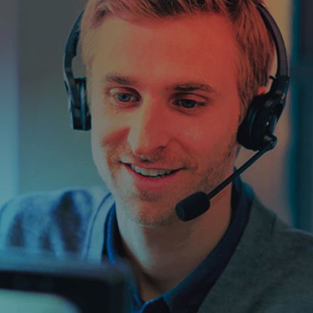 A person wearing a headset is smiling while looking at a computer screen. They appear to be engaged in a conversation or providing assistance, with a warm blue and orange lighting in the background.