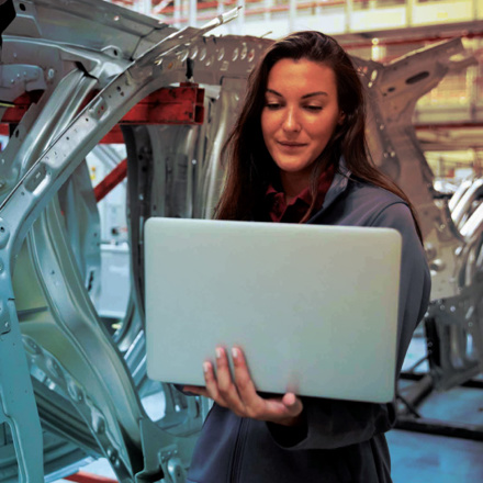 A woman in a gray jacket stands in an industrial setting, holding a laptop. Behind her are metal car frames, suggesting a manufacturing environment. She appears focused on her work.