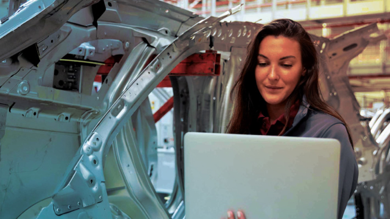 A woman in a gray jacket stands in an industrial setting, holding a laptop. Behind her are metal car frames, suggesting a manufacturing environment. She appears focused on her work.