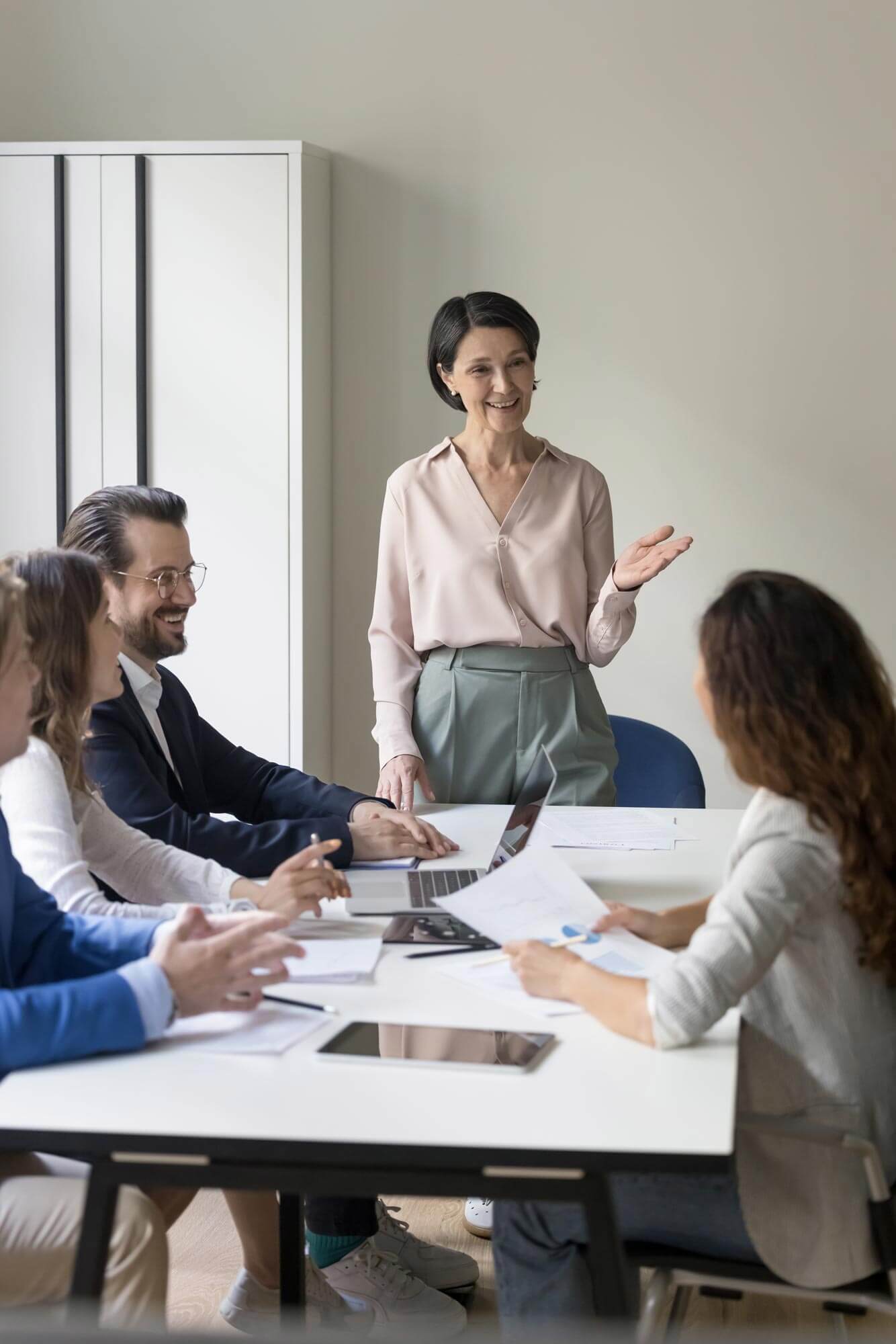 Business lady or coach lead group meeting or training in conference room. Clients take part in negotiations provide by mature company boss joking, provide information solve business laugh during break.