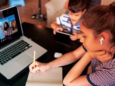 A girl with earbuds writes in a notebook while looking at a laptop screen, possibly participating in an online class. In the background, a boy uses a tablet. They are in a cozy indoor setting.