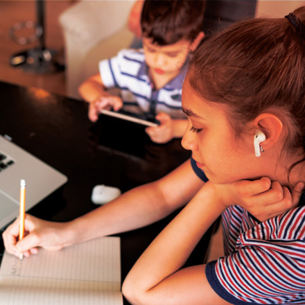 A girl with earbuds writes in a notebook while looking at a laptop screen, possibly participating in an online class. In the background, a boy uses a tablet. They are in a cozy indoor setting.