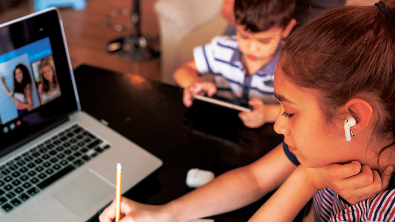 A girl with earbuds writes in a notebook while looking at a laptop screen, possibly participating in an online class. In the background, a boy uses a tablet. They are in a cozy indoor setting.