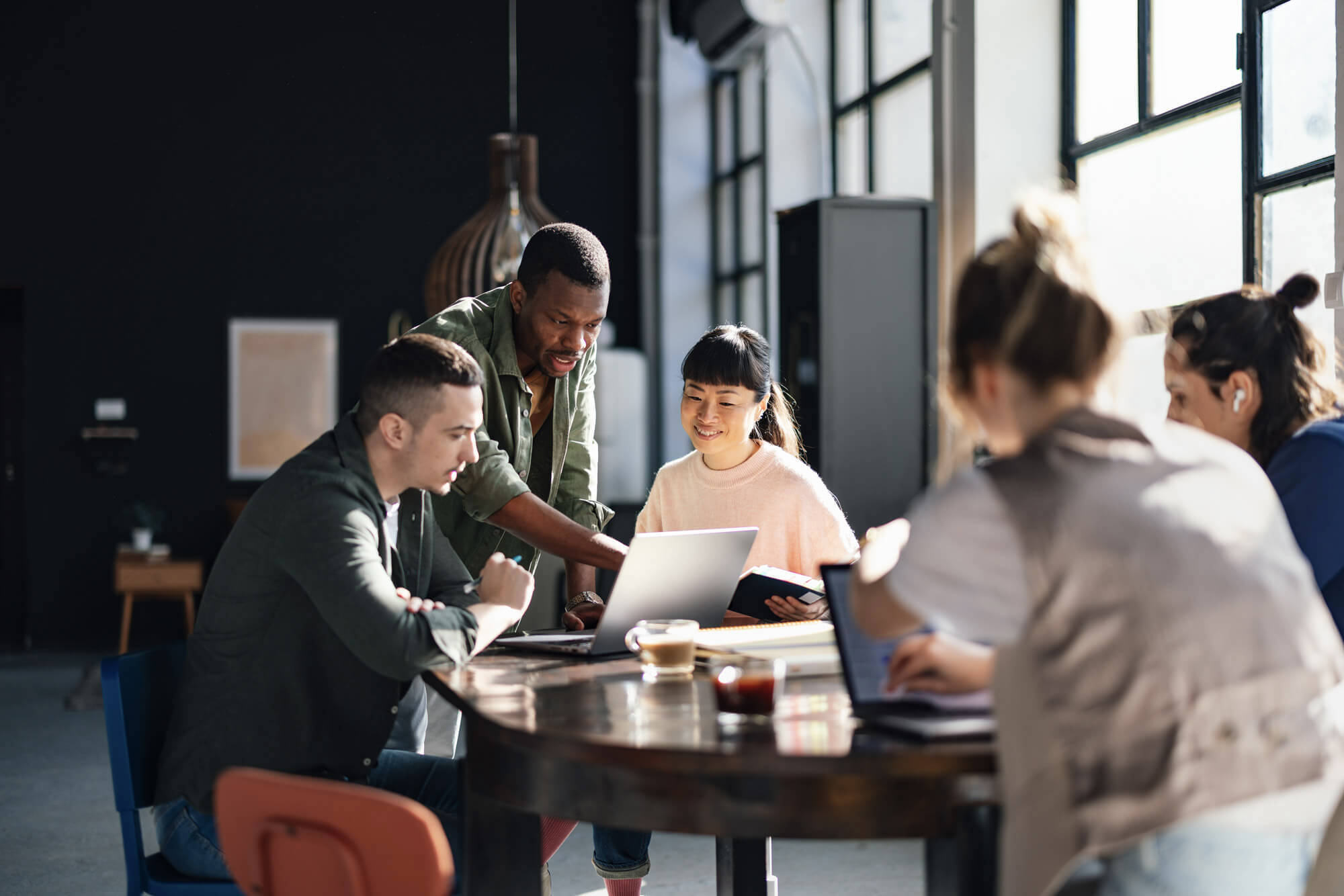 A diverse group of four people are gathered around a wooden table, collaborating with a laptop in a modern workspace. One person is pointing at the screen while others are actively engaged. Large windows in the background bring in natural light.