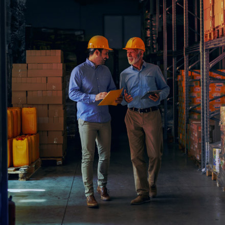 Two business partners in formal wear and with protective yellow helmets on heads walking and talking about business. Younger one holding folder with data while older one using tablet.