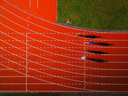 Aerial view of four runners sprinting on a red athletics track. Their shadows stretch to the right, and the green grass outside the track borders the image at the top. White lane markings are clearly visible.