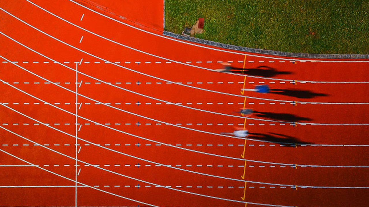 Aerial view of four runners sprinting on a red athletics track. Their shadows stretch to the right, and the green grass outside the track borders the image at the top. White lane markings are clearly visible.