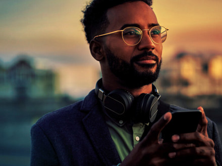 A bearded man wearing glasses and headphones is holding a smartphone. He stands outdoors at sunset, looking thoughtfully into the distance. The background is softly blurred, highlighting the warm glow of the sky.