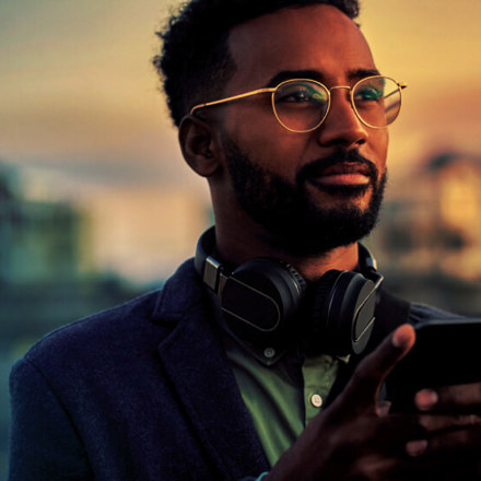 A bearded man wearing glasses and headphones is holding a smartphone. He stands outdoors at sunset, looking thoughtfully into the distance. The background is softly blurred, highlighting the warm glow of the sky.