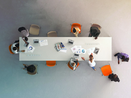 Aerial view of a conference table with ten people seated around it. They are discussing documents and using laptops. The table is white, and four orange chairs stand out among others, suggesting a meeting or collaboration.