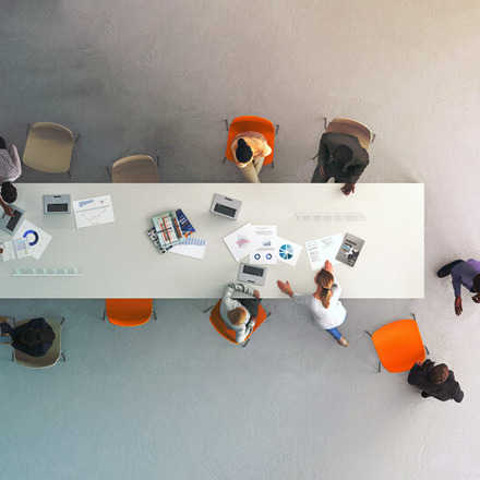 Aerial view of a conference table with ten people seated around it. They are discussing documents and using laptops. The table is white, and four orange chairs stand out among others, suggesting a meeting or collaboration.