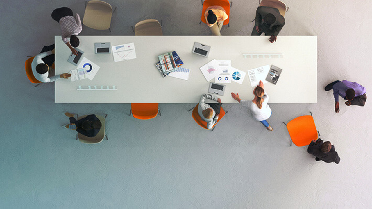 Aerial view of a conference table with ten people seated around it. They are discussing documents and using laptops. The table is white, and four orange chairs stand out among others, suggesting a meeting or collaboration.