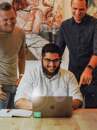 Four people are gathered around a table, smiling and looking at a laptop screen. One person is seated, using the laptop, while the others stand or sit nearby. There's artwork on the wall behind them.