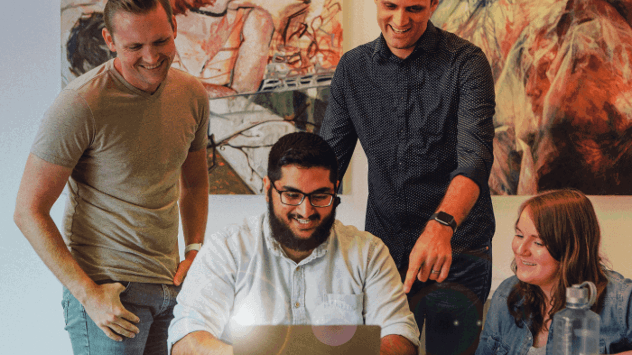 Four people are gathered around a table, smiling and looking at a laptop screen. One person is seated, using the laptop, while the others stand or sit nearby. There's artwork on the wall behind them.