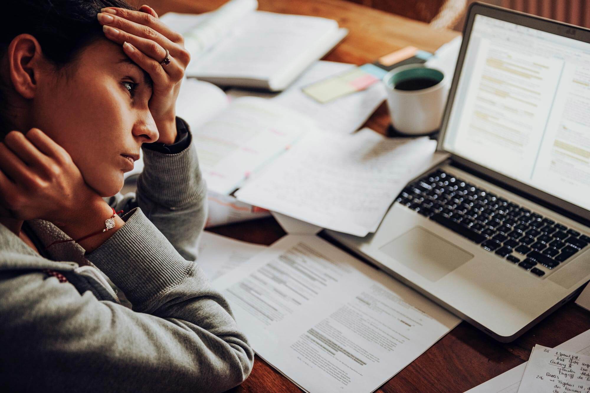 A person looking stressed sits at a table surrounded by papers and an open laptop displaying documents. They rest their head on one hand, with a cup of coffee nearby, suggesting they are studying or working intensely.