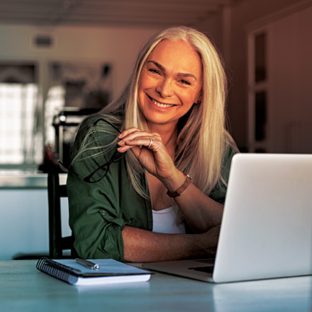 A smiling woman with long gray hair sits at a desk holding glasses in one hand. She is in a well-lit room with a notebook, pen, and laptop in front of her. She wears a green shirt over a white top.