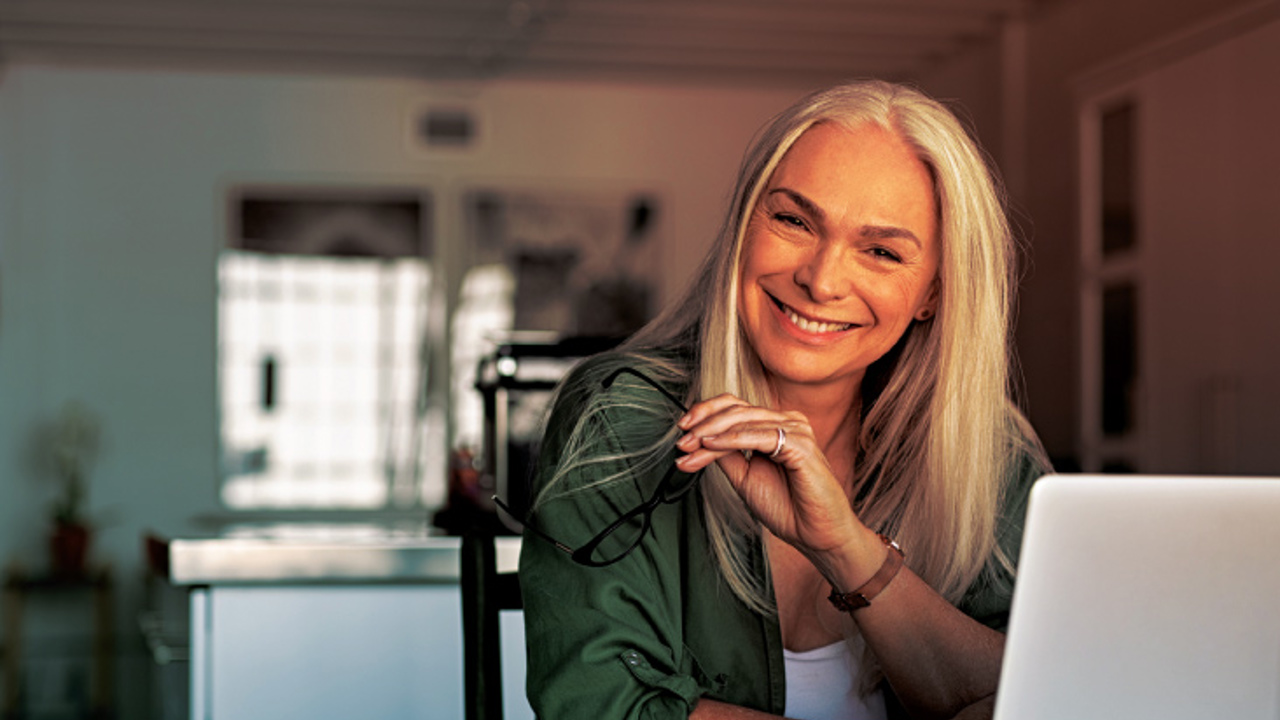 A smiling woman with long gray hair sits at a desk holding glasses in one hand. She is in a well-lit room with a notebook, pen, and laptop in front of her. She wears a green shirt over a white top.