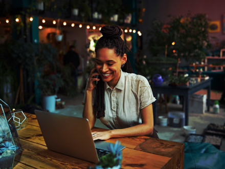 Smiling pleased pretty female florist with cornrows sitting at the laptop during a phone conversation