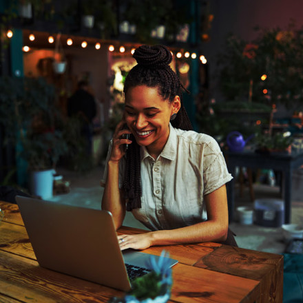 Smiling pleased pretty female florist with cornrows sitting at the laptop during a phone conversation