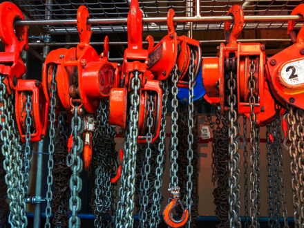 A close-up of several red manual chain hoists hanging vertically. The hoists have chains dangling below and are arranged in a row against a metal grate background. One hoist is labeled with a '2 ton' capacity sign.