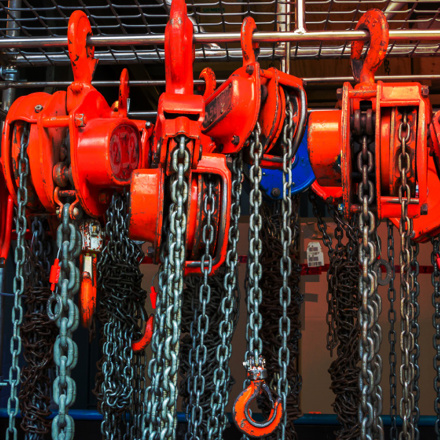 A close-up of several red manual chain hoists hanging vertically. The hoists have chains dangling below and are arranged in a row against a metal grate background. One hoist is labeled with a '2 ton' capacity sign.