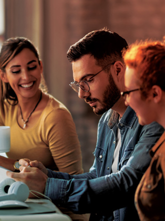 Three people sit at a desk, collaborating on a project. A man in the middle reviews a document, while the women on either side of him smile. The desk has a laptop, headphones, and a coffee mug. The setting is lit warmly, suggesting a casual work environment.