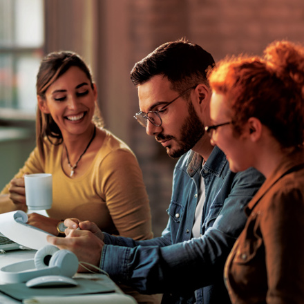 Three people sit at a desk, collaborating on a project. A man in the middle reviews a document, while the women on either side of him smile. The desk has a laptop, headphones, and a coffee mug. The setting is lit warmly, suggesting a casual work environment.