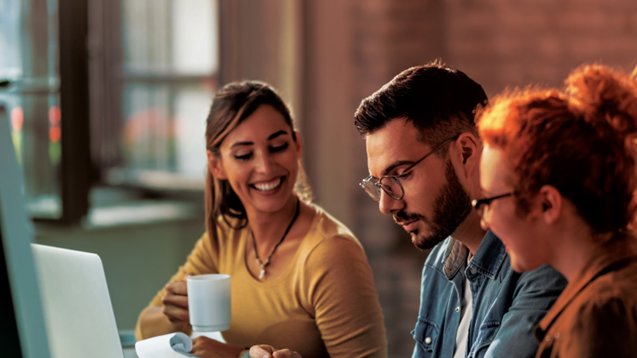 Three people sit at a desk, collaborating on a project. A man in the middle reviews a document, while the women on either side of him smile. The desk has a laptop, headphones, and a coffee mug. The setting is lit warmly, suggesting a casual work environment.