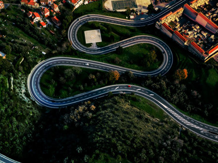 Aerial view of a winding road with sharp curves cutting through a green landscape. A red and a white car are on the road. Nearby, there are residential houses and a basketball court visible in the top part of the image.