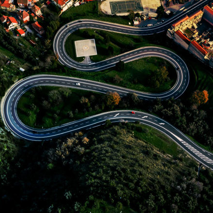 Aerial view of a winding road with sharp curves cutting through a green landscape. A red and a white car are on the road. Nearby, there are residential houses and a basketball court visible in the top part of the image.