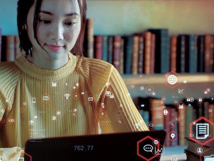 A woman in a yellow sweater works on a laptop in a library. Digital icons float around her, symbolizing connectivity and data. Bookshelves are visible in the background.