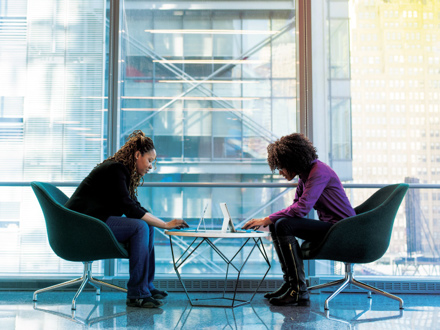 Two people sit across from each other at a small table in a modern office setting. Both are focused on their laptops. Large windows in the background reveal an urban cityscape. The chairs are sleek and contemporary.