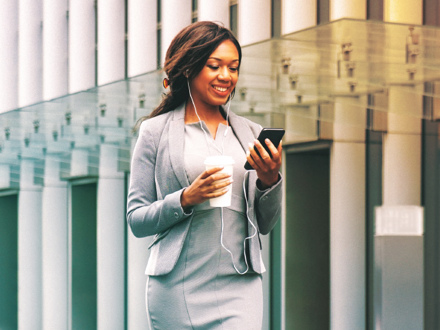 A woman in a business suit smiles while looking at her smartphone. She wears earphones and holds a takeaway coffee cup. The background features modern glass architecture, suggesting an urban environment.