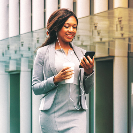 A woman in a business suit smiles while looking at her smartphone. She wears earphones and holds a takeaway coffee cup. The background features modern glass architecture, suggesting an urban environment.