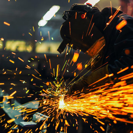 A person wearing protective gear operates a welding tool, sending a shower of bright orange sparks through the air in a dimly lit workshop.