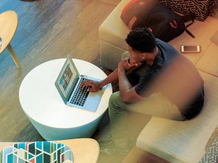 A person sitting on a beige couch using a laptop on a round white table. A backpack and a smartphone are beside them. The setting appears to be indoors with wooden flooring and modern furniture.