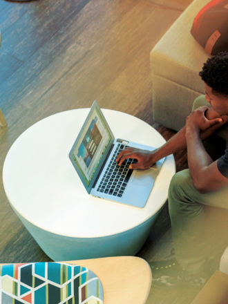 A person sitting on a beige couch using a laptop on a round white table. A backpack and a smartphone are beside them. The setting appears to be indoors with wooden flooring and modern furniture.