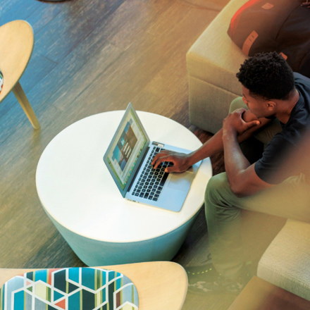 A person sitting on a beige couch using a laptop on a round white table. A backpack and a smartphone are beside them. The setting appears to be indoors with wooden flooring and modern furniture.