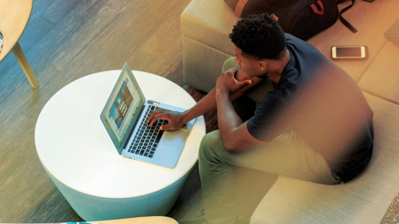 A person sitting on a beige couch using a laptop on a round white table. A backpack and a smartphone are beside them. The setting appears to be indoors with wooden flooring and modern furniture.