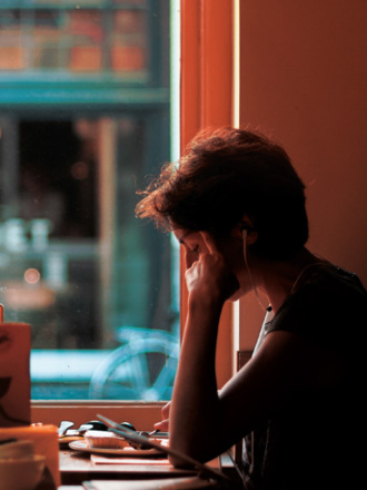 A person wearing headphones sits by a window in a dimly lit café, resting their head on their hand. The café window shows blurred outdoor scenery with a bicycle and the word 'CABINET' visible in the background.