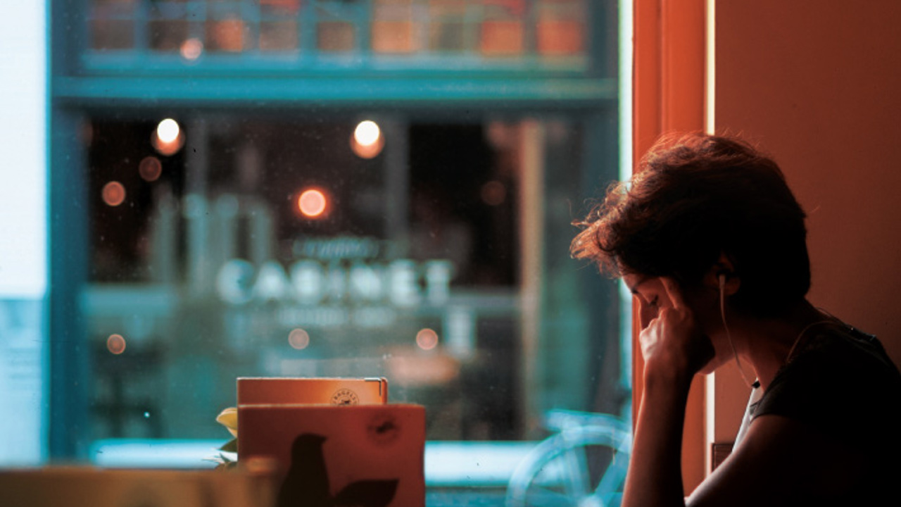 A person wearing headphones sits by a window in a dimly lit café, resting their head on their hand. The café window shows blurred outdoor scenery with a bicycle and the word 'CABINET' visible in the background.