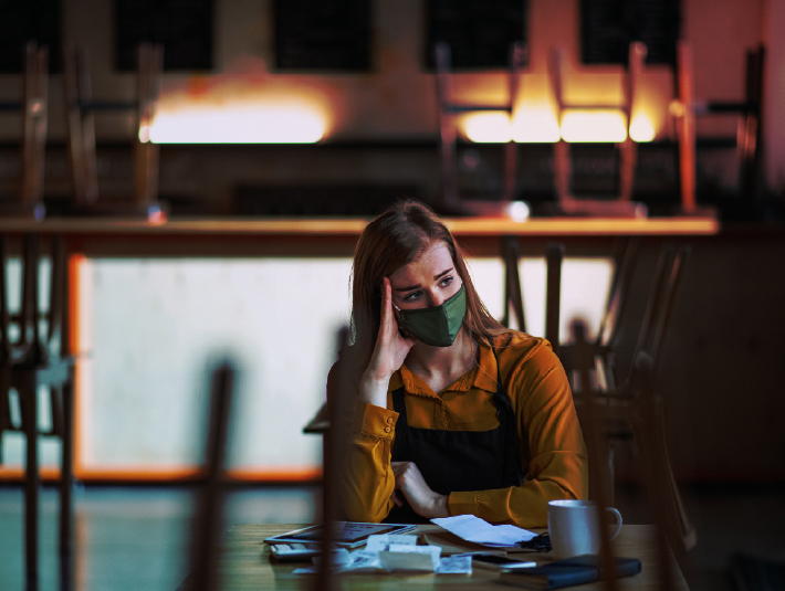 A woman wearing a green mask and orange shirt sits at a table with papers and a cup, resting her head on her hand. The chairs in the background are stacked on tables, indicating a closed or empty restaurant.