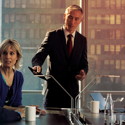 Three people are in an office meeting. A man in a suit is standing, pointing at a tablet, while a woman and another man sit at a table with coffee mugs and laptops. Sunlight filters through a large window behind them.